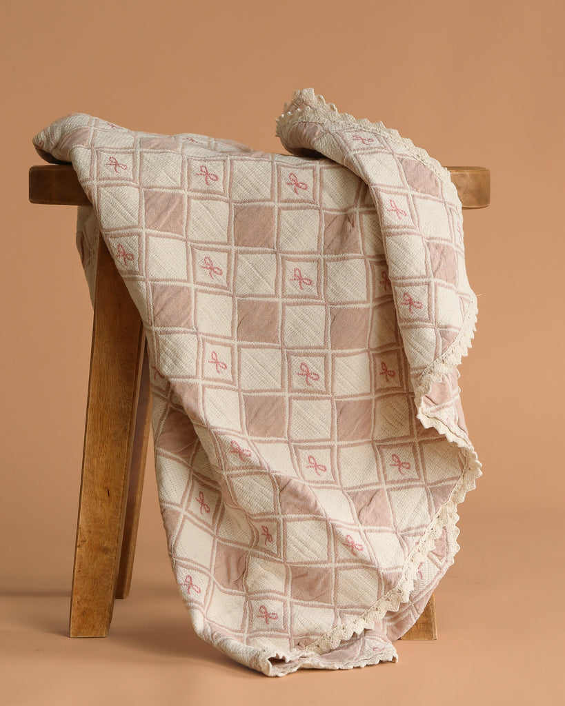 The Organic Cotton Baby Patchwork Blanket - Pink Bows features soft beige and pink embroidery with lace edging, making it perfect as a cozy baby blanket or nursery accent.