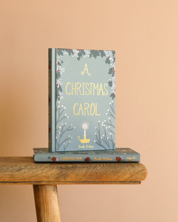 A Christmas Carol | Collector's Edition| Hardcover Book with a blue-green floral cover stands upright on a wooden bench over another copy, set against a beige background, capturing the timeless spirit of Dickens' classic tale.