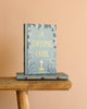 A Christmas Carol | Collector's Edition| Hardcover Book with a blue-green floral cover stands upright on a wooden bench over another copy, set against a beige background, capturing the timeless spirit of Dickens' classic tale.