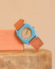 The Mini Kyomo Watch - Havana Sky, with a blue face and orange strap, rests on a peach block atop a wooden surface against a beige background.