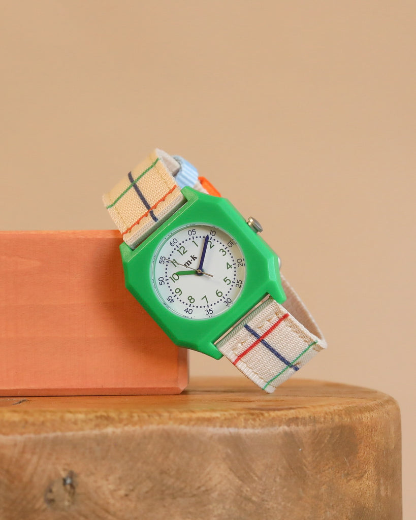 The Mini Kyomo Watch x Taxi Brousse - Color Notebook, with a green octagonal case and colorful plaid strap, sits on an orange block atop wood in a playful vintage style against a beige background.