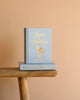 A light blue hardcover "Sense and Sensibility | Collector's Edition" with gold lettering and two hats is displayed on a rustic wooden bench against a peach background.
