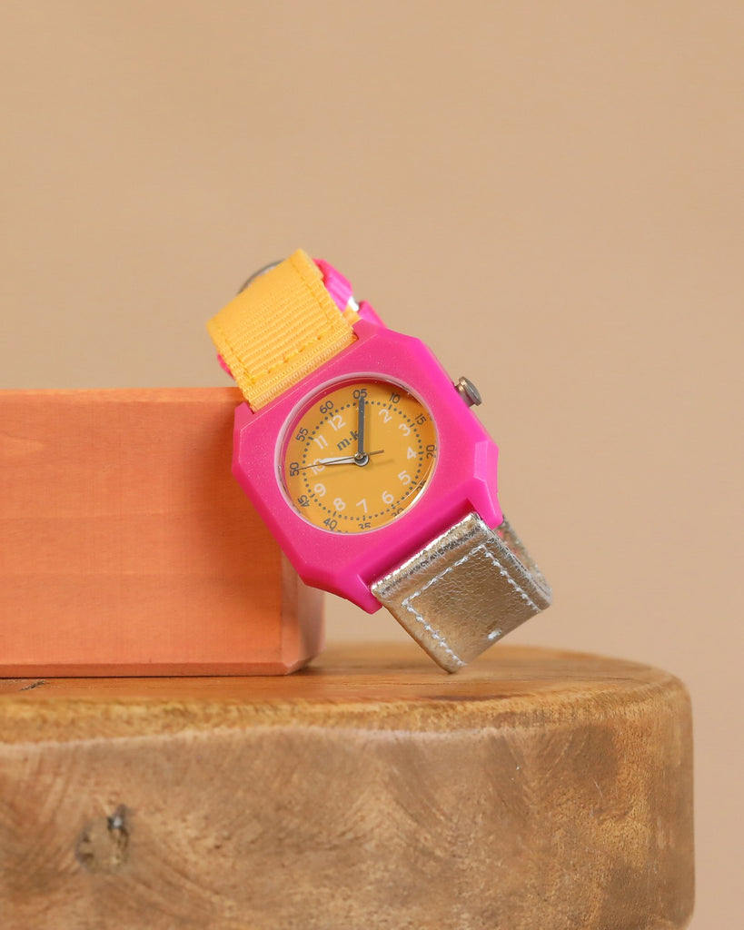 The Mini Kyomo Watch - Disco, featuring a pink case, yellow face and band, and a silver strap end, is displayed on an orange block atop a round wooden surface with a beige background.