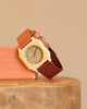 A Mini Kyomo Watch - Cherry Bomb with a beige octagonal face and brown fabric strap rests on wood, leaning against a peach block with a neutral beige background.