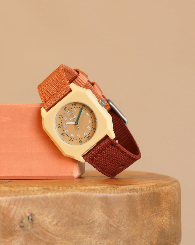 A Mini Kyomo Watch - Cherry Bomb with a beige octagonal face and brown fabric strap rests on wood, leaning against a peach block with a neutral beige background.