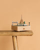 The Paris Music Box, adorned with miniatures of the Eiffel Tower, a bridge, trees, and a boat, is displayed on a rustic wooden stool set against a plain beige background.