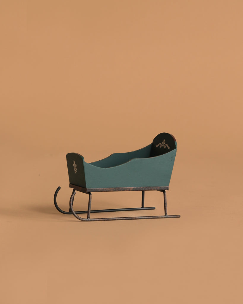 The Maileg Sleigh, Mouse - Green is a small, vintage-style green wooden sleigh with black runners and white hand-painted motifs, displayed on a plain light brown background.