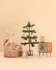 A Maileg Cozy Christmas Corner with Little Sister mouse in a pink dress sits in a plush armchair next to a miniature Christmas tree, a small stool, and a vintage-style television topped with a yellow alarm clock.