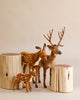 A group of Large Deer Stuffed Animal - Male (final sale), including a stag with antlers, a doe, and a fawn, stand on a light-colored background. Featuring hand-sewn details, they are positioned around two large, smooth tree stump props. The deer toys have brown fur with white spots on their backs.