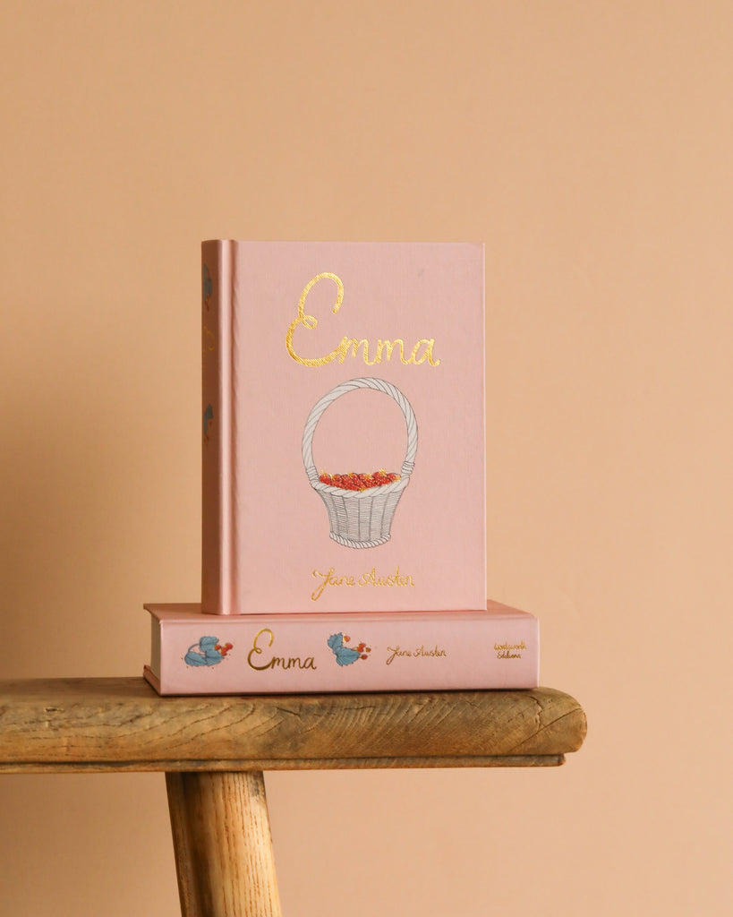 The "Emma | Collector's Edition | Hardcover" in pink, featuring a white basket illustration, stands upright on a wooden bench with another matching copy lying flat beneath it against a peach-colored background.