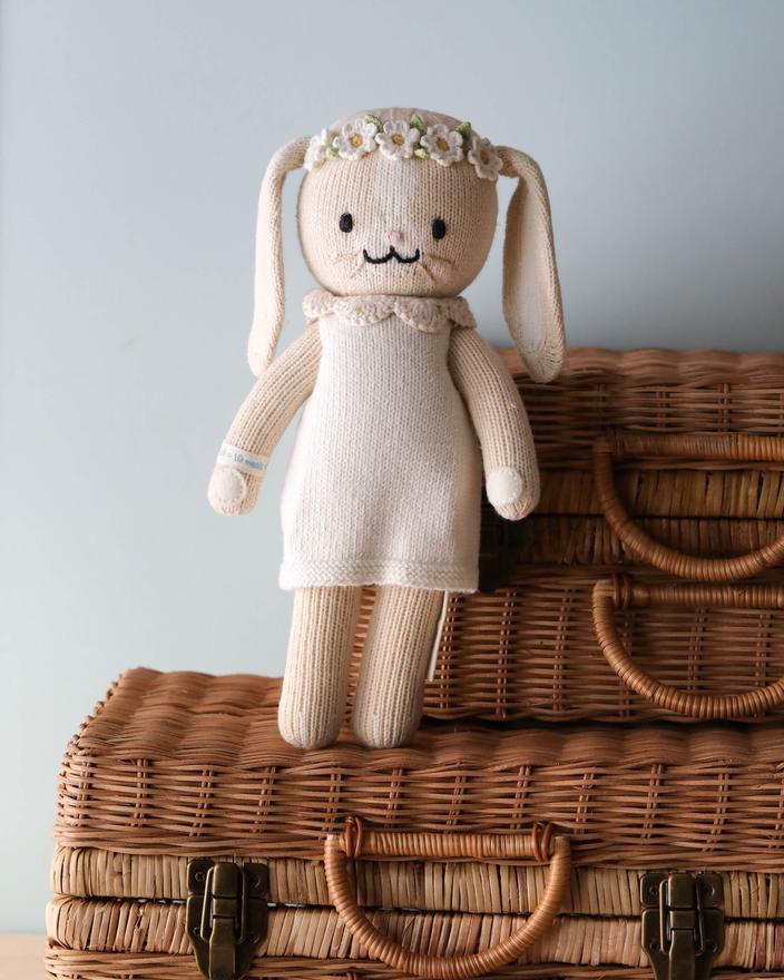 Cuddle + Kind Hannah the Bunny, hand-knit from premium cotton yarn and dressed in a cream dress with a flower crown, sits on two stacked wicker suitcases against a light blue wall.