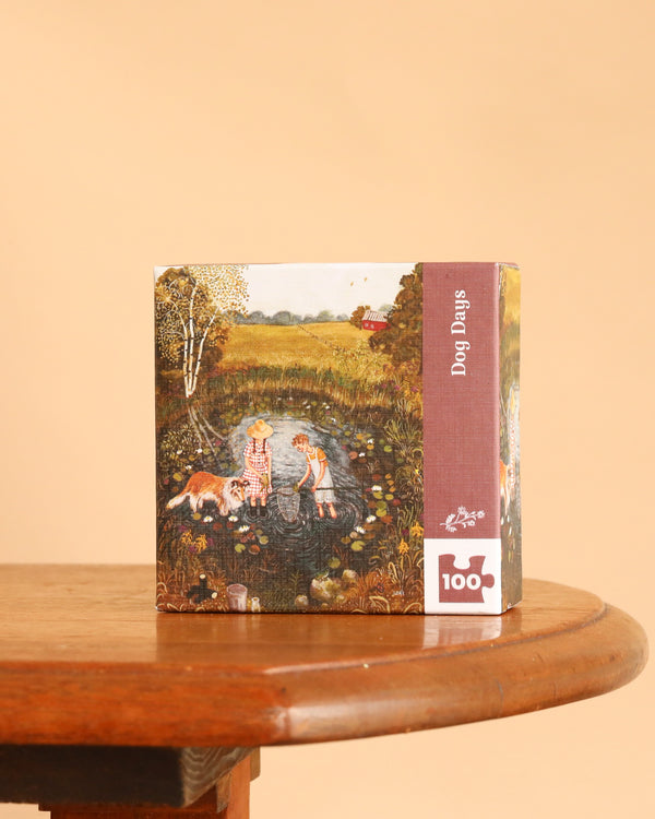 A square Dog Days Mini - 100 Piece Mini Puzzle box sits on a wooden table, featuring an illustration of two children and a dog at a pond, with the title and piece count printed on the side.