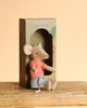 A Maileg Little Green Thumb Garden Set mouse sits on a wooden surface in front of a "mimie" box, wearing a red and white striped shirt and blue skirt, with a beige background behind.