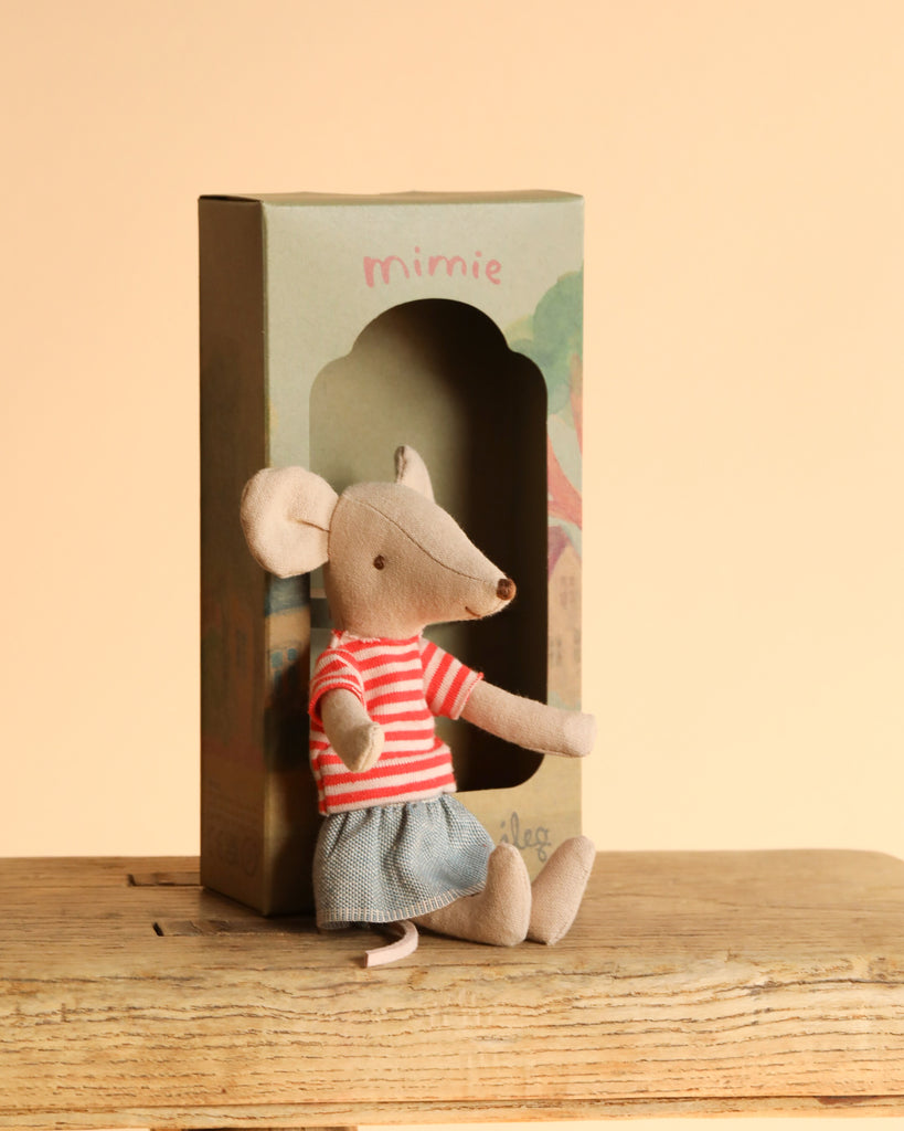A Maileg Little Green Thumb Garden Set mouse sits on a wooden surface in front of a "mimie" box, wearing a red and white striped shirt and blue skirt, with a beige background behind.