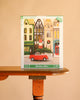 Christmas Cheer - 1000 Piece Jigsaw Puzzle box featuring a Janet Hill illustration of a red car carrying a Christmas tree through a city street lined with shops.