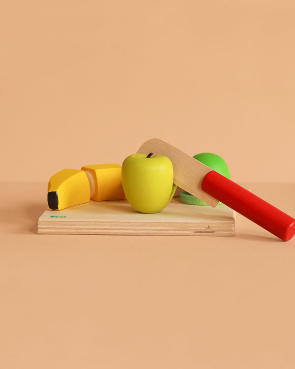 The Erzi Fruit Salad Cutting Set includes a yellow banana, green apple, and red-handled knife on a small cutting board—perfect wooden fruit toys for pretend play kitchen fun against a beige background.