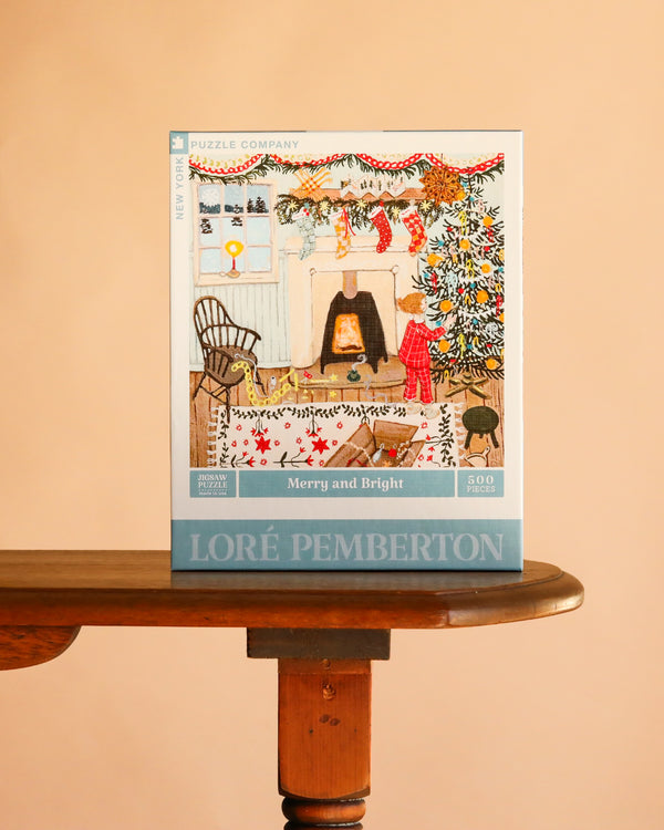 A Merry and Bright - 500 Piece Jigsaw Puzzle box showing an illustration of a child decorating a tree by a fireplace, resting on a wooden table.