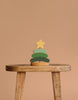The Handmade Wooden Stacking Toy Green Christmas Tree, featuring green layers and a yellow star topper, is displayed on a rustic wooden stool against a plain beige background.
