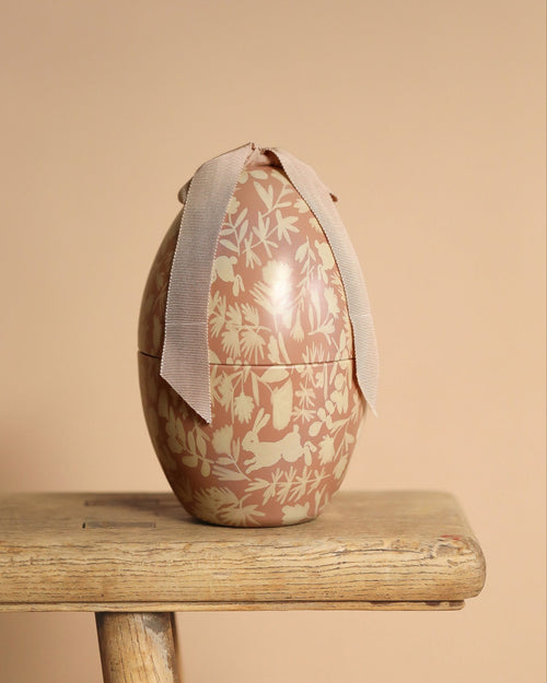 A Maileg Easter Egg, Medium - Powder with a cream floral and bunny pattern sits on a wooden stool, tied with a light pink ribbon and standing out beautifully against the beige background.