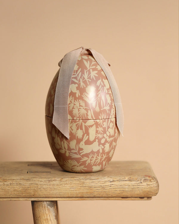A Maileg Easter Egg, Medium - Powder with a cream floral and bunny pattern sits on a wooden stool, tied with a light pink ribbon and standing out beautifully against the beige background.