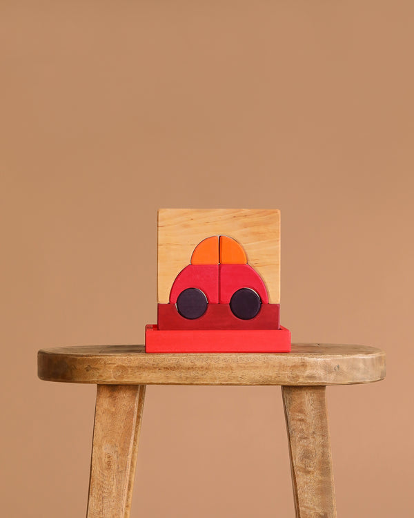 The Grimm's Building Set - Red, a wooden puzzle resembling a red car with black wheels and orange windows, is displayed assembled on a rustic stool—perfect for any wooden toy or color block collection—against a plain beige background.