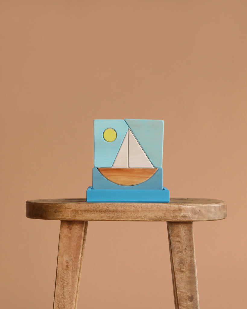 The Grimm's Building Set - Blue, a wooden stool topped with a colorful geometric block puzzle featuring a sailboat and sun, highlights classic toddler toys made from wooden building blocks against a beige background.