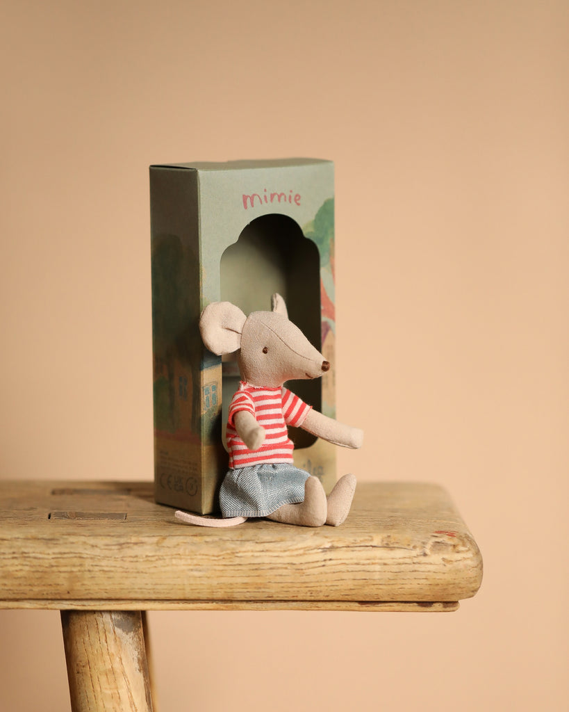 A Maileg Big Sister and Me Nursery Set features a small mouse toy dressed in a red striped shirt and blue shorts, seated on a wooden stool with its box visible in the background against a plain beige backdrop.