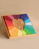 A Grimm's Rainbow Lion Building Set featuring a lion's face in the center. Surrounding the lion's head are colorful geometric shapes arranged in a radial pattern, painted in vibrant gradient colors ranging from reds and oranges to blues and greens. The puzzle sits in a natural wood tray.