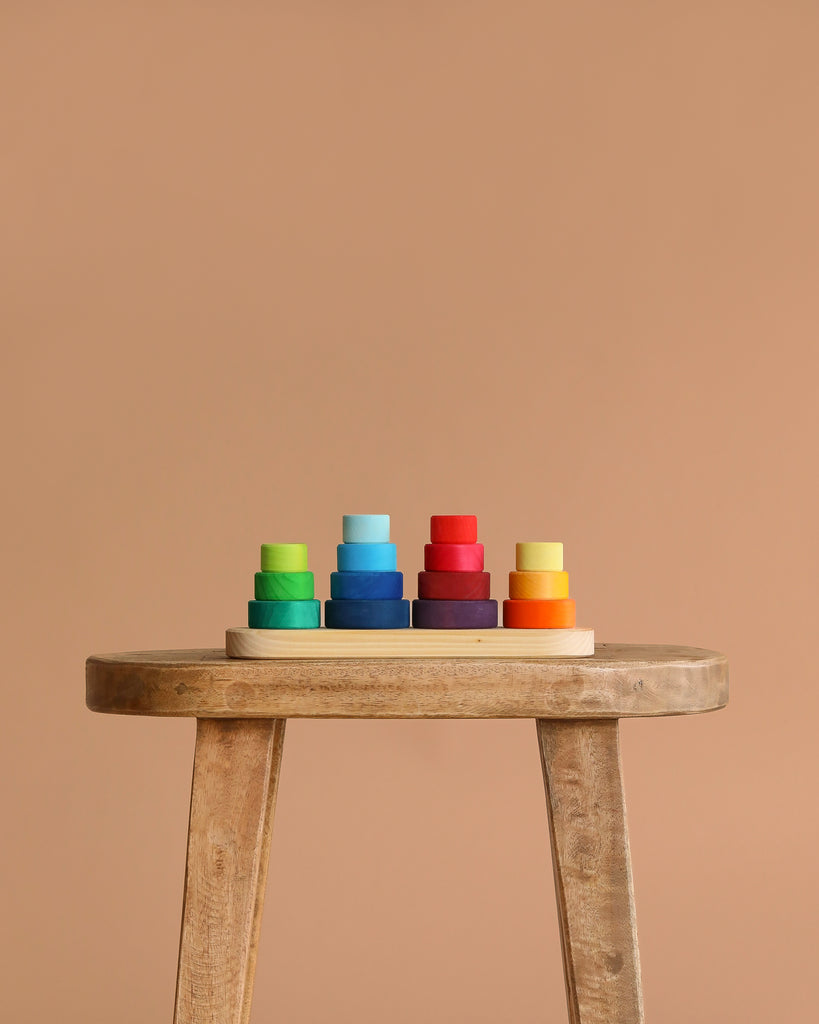 Grimm's Fabuto Stacker, a colorful wooden stacking toy with green, blue, red, yellow, and orange rings, is neatly arranged on a light wooden stool against a beige background.
