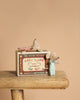 Two small mice from the Maileg Family Celebration Set, one in pajamas and one snuggled under a polka dot blanket, are posed with their “Baby Twins Safety Matches” box on a wooden stool against a plain beige background—an adorable set perfect for collectors and imaginative play.