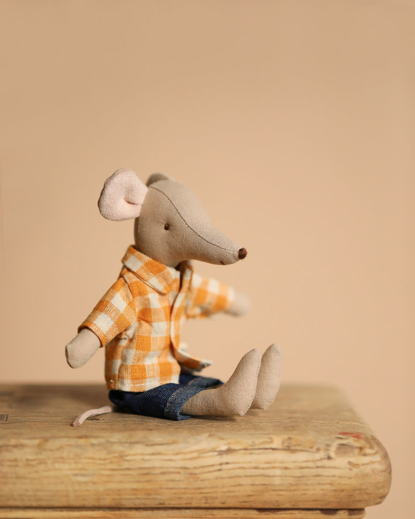The Maileg Dad Mouse with an extra outfit, dressed in a yellow and white checkered shirt and blue pants, sits on a wooden surface, exuding endearing style against a warm beige background.