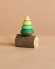 The Mader Spinning Tree Top On Trunk, a small green pine tree toy crafted from Maple wood, is stacked on a short cylindrical piece of tree branch against a plain beige background.