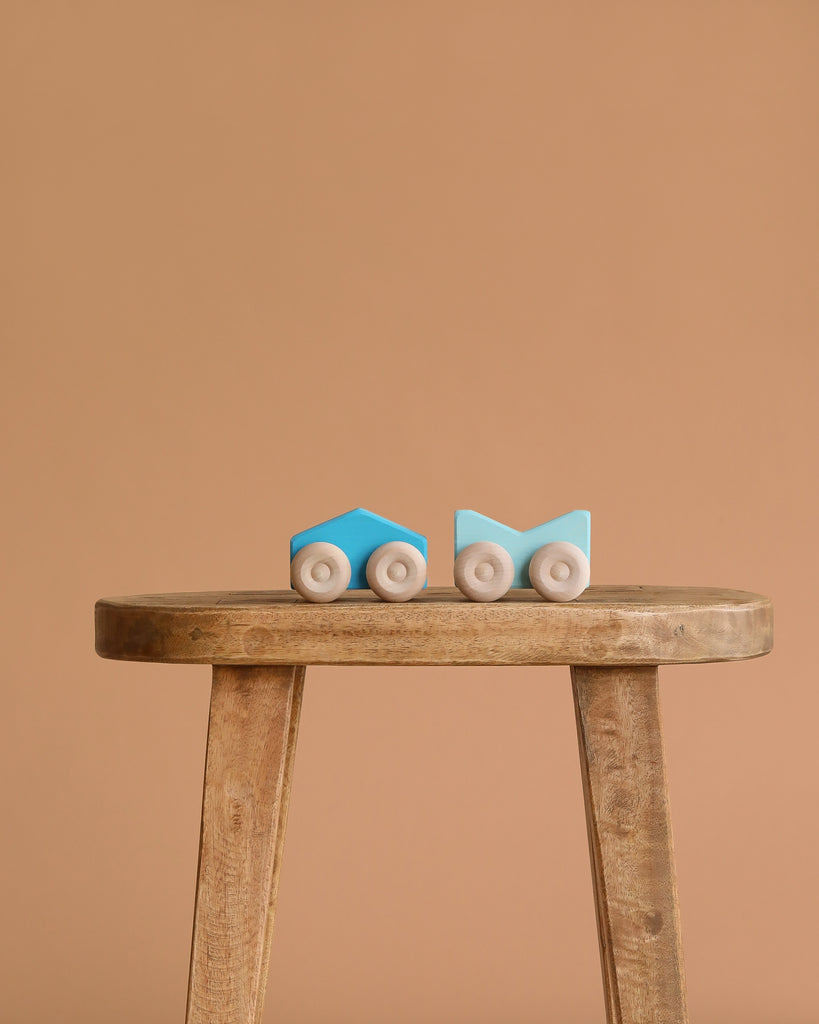 Two small Grimm's Stacking Cars - Blue, featuring minimalist designs, sit side by side on a wooden stool against a plain light brown background.