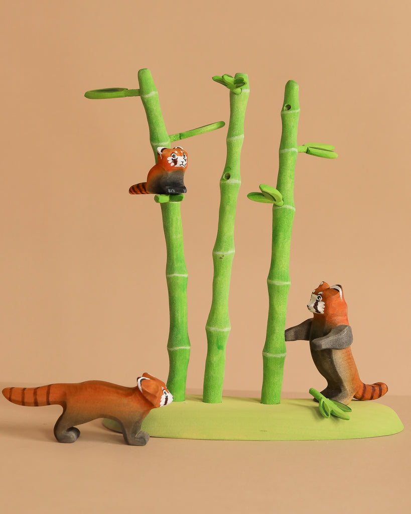 Three small, handcrafted figures from the Bumbu Handmade Wooden Red Panda Family with Bamboo Set interact on a green base, with one red panda climbing bamboo, another standing upright, and the third walking nearby against a beige backdrop.