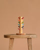 The Grimm's Sorting Helper with Building Rings Set features colorful stacking pegs arranged in pairs, displayed upright on a rustic wooden stool against a plain beige background—ideal for developing fine motor skills.