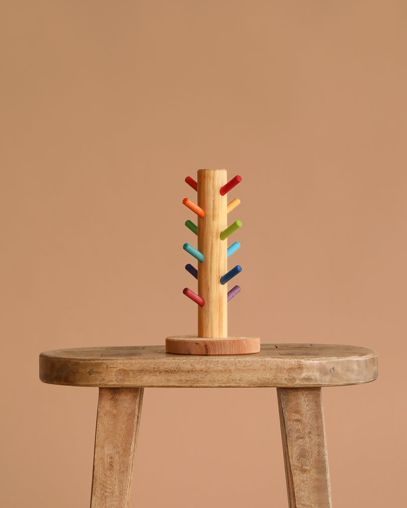 The Grimm's Sorting Helper with Building Rings Set features colorful stacking pegs arranged in pairs, displayed upright on a rustic wooden stool against a plain beige background—ideal for developing fine motor skills.