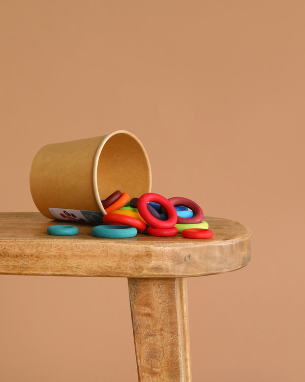 A brown paper cup tipped over on a wooden surface, spilling Grimm's Building Rings - Rainbow from a stacking toy onto the table against a plain beige background—ideal for developing fine motor skills.