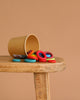 A brown paper cup tipped over on a wooden surface, spilling Grimm's Building Rings - Rainbow from a stacking toy onto the table against a plain beige background—ideal for developing fine motor skills.