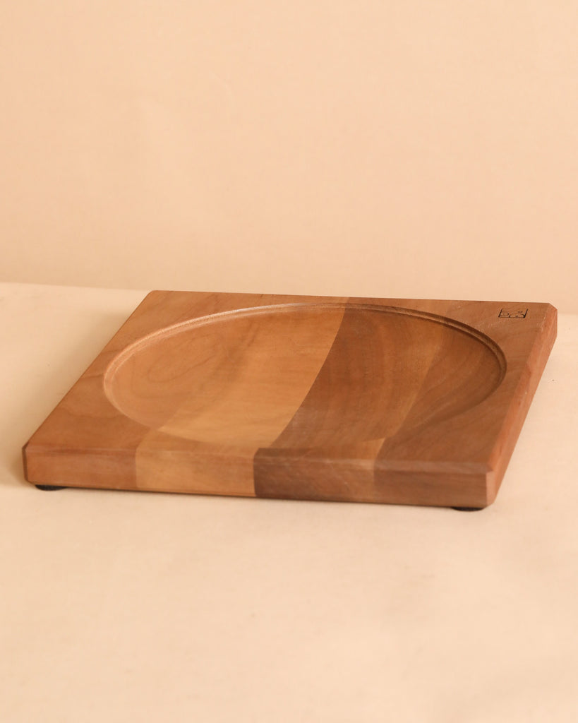 The Walnut Wood Spinning Plate is a square plate with a central round, shallow indent and natural wood grain, crafted to meet toy safety standards. It is displayed on a light beige surface with a matching background.