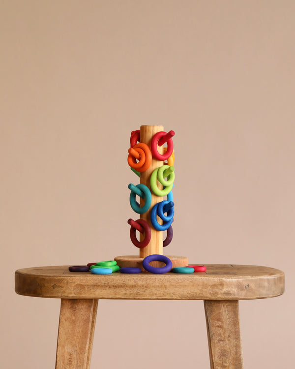 The Grimm's Sorting Helper with Building Rings Set, featuring colorful rings, sits on a wooden stool against a beige background—ideal for developing fine motor skills as some rings are stacked while others are scattered.