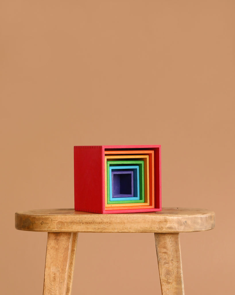 Grimm's Large Set of Boxes - Rainbow is displayed stacked by size and color on a wooden stool against a beige background, with the largest box in red and the smallest dark blue at the center.