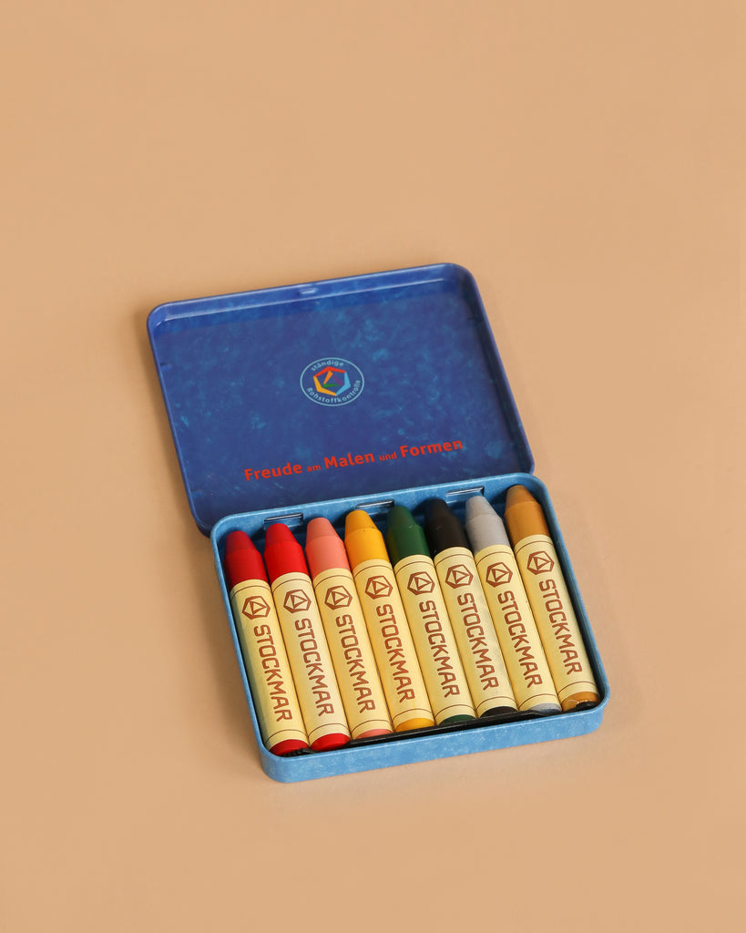 The Stockmar Wax Stick Crayons Tin Case - 8 Supplementary Colors includes eight non-toxic crayons in assorted shades, neatly arranged side by side in a blue tin—perfect for art education.