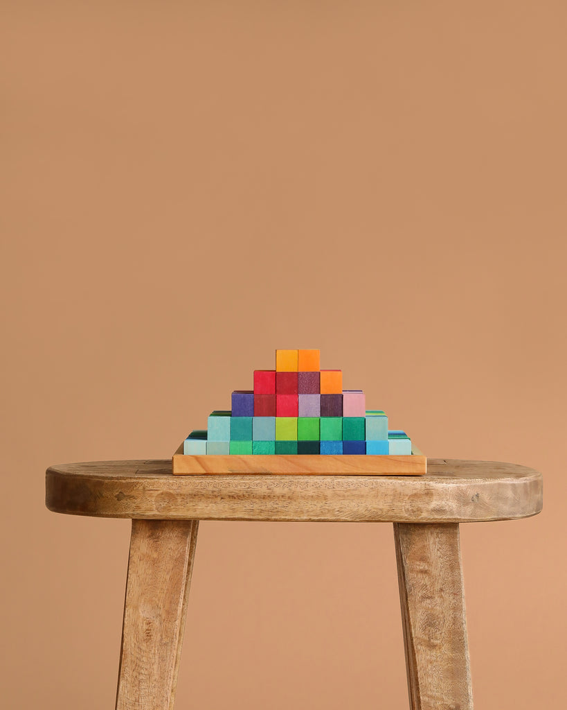 A wooden stool displays a neatly stacked pyramid of Grimm's Stepped Pyramid Block Set - Small against a plain beige background.
