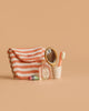 A striped pink and white pouch sits beside a Maileg Bathroom Set, a small bottle labeled "Flora," pink lipstick, a gold-framed handheld mirror, and a pink toothbrush in a white cup on a beige background.