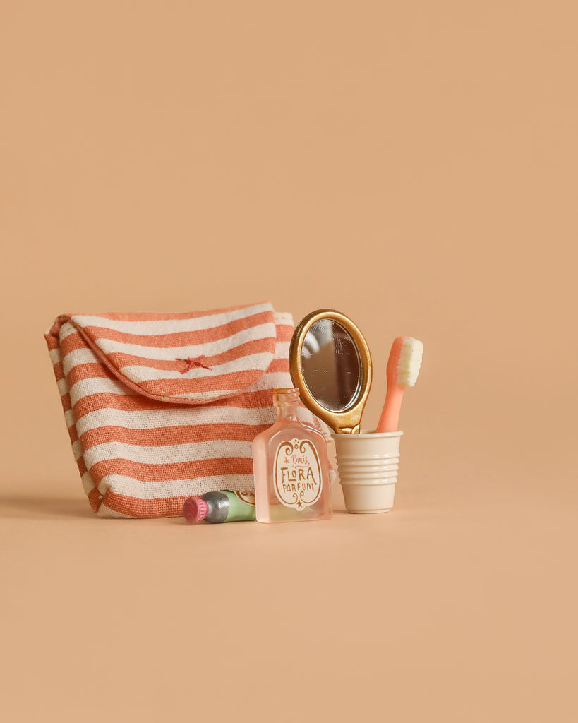 A striped pink and white pouch sits beside a Maileg Bathroom Set, a small bottle labeled "Flora," pink lipstick, a gold-framed handheld mirror, and a pink toothbrush in a white cup on a beige background.