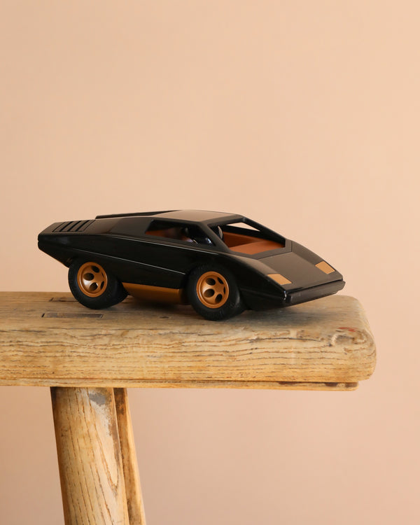 The Playforeer UFO Cannone Car in Black, styled after 70's concept cars with gold accents, is displayed on a rustic wooden stool against a plain beige background.