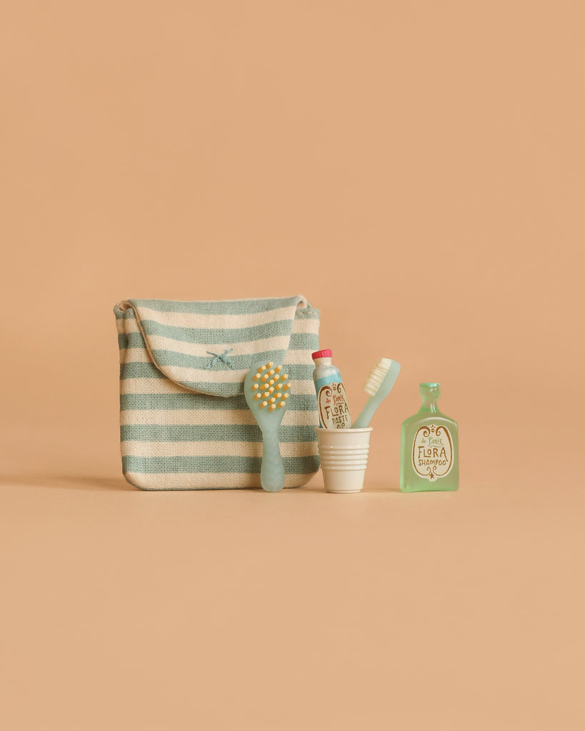 The Maileg Toothbrush Set in Bag, Mouse - Blue Stripe features a striped pouch, tiny hairbrush, cup with toothbrush and toothpaste, and a mini "FLORA" bottle, all displayed against a plain beige background.