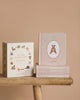 A set of baby board books from the Our Little Library Book Box Set rests on a wooden bench against a beige background. Perfect for speech therapists, it includes themed titles like 'First Words,' 'Transportation,' 'Fruits & Veggies,' 'Forest Animals,' and 'Farm Animals.'.