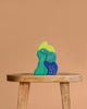 Grimm's Casa Aqua Building Blocks—abstract, colorful pieces in green, blue, and teal with white line drawings—are arranged on a wooden stool against a plain beige background. Their irregular shapes inspire imaginative play.