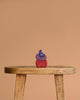 A trio of Grimm's Small Bakery Building Blocks—blue, purple, and red—are stacked atop a rustic wooden stool, standing out against a plain beige background.